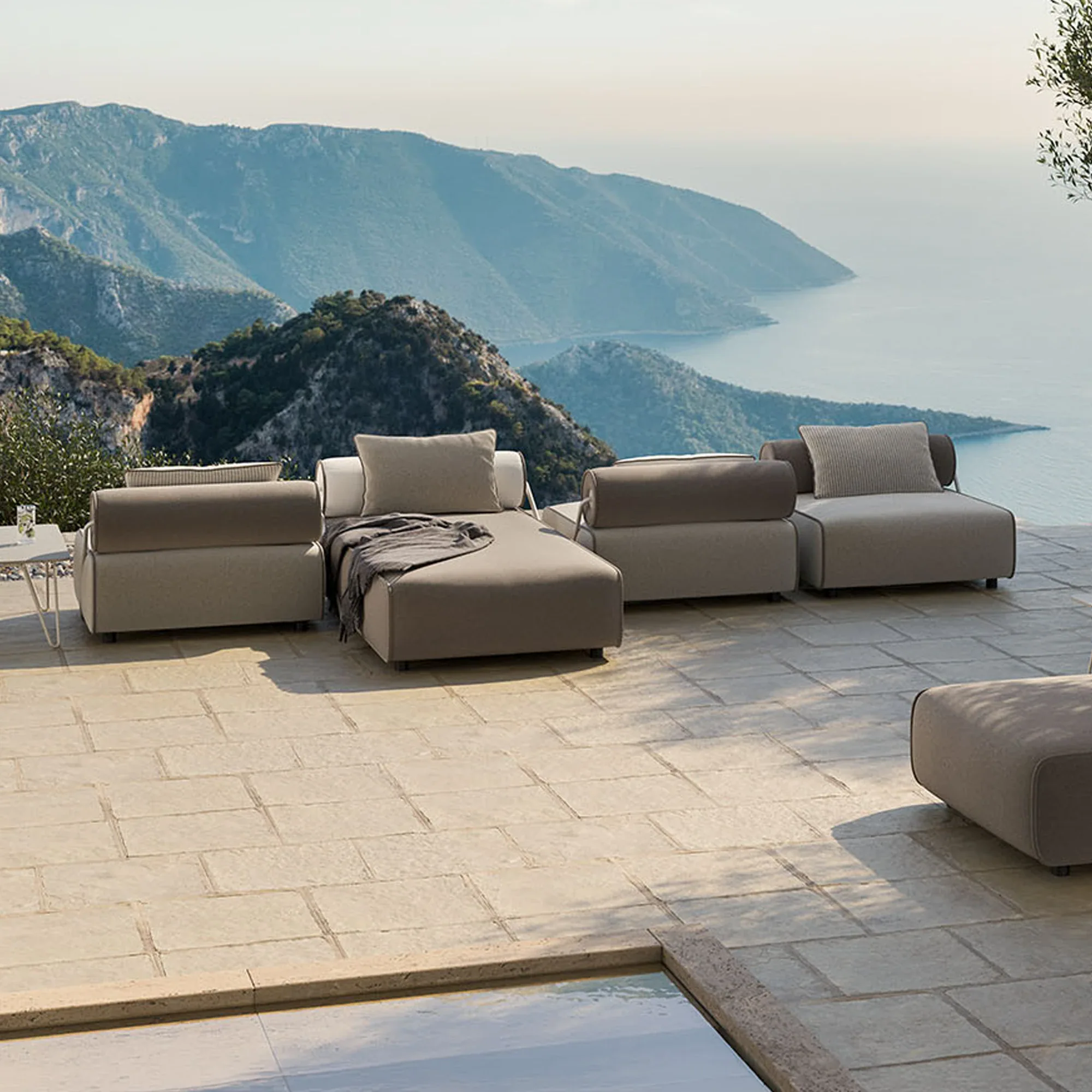 An outdoor patio with modern modular beige and brown seating, including sofas and a chaise lounge, overlooking a scenic vista of green mountains and a vast blue sea under a clear sky.