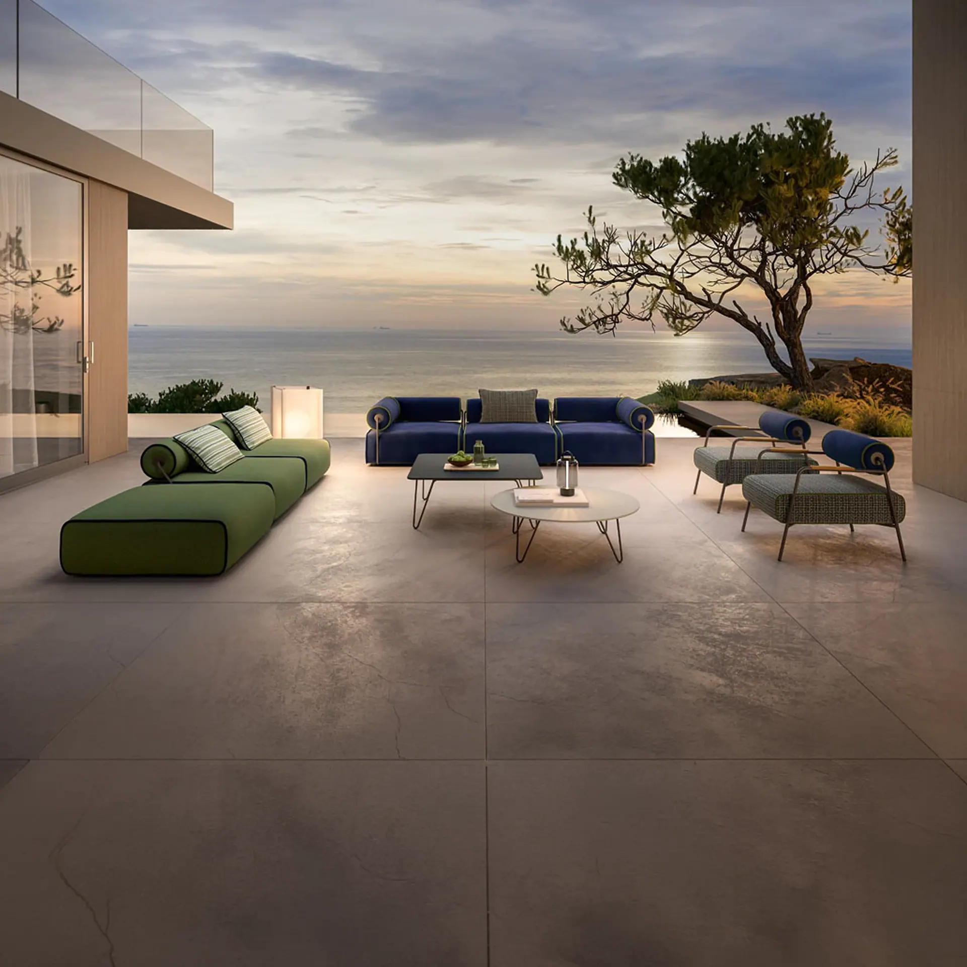 An elegant outdoor patio with modern green and blue furniture, coffee tables, and lanterns, overlooking a calm ocean at sunset with a gnarled tree on the right.
