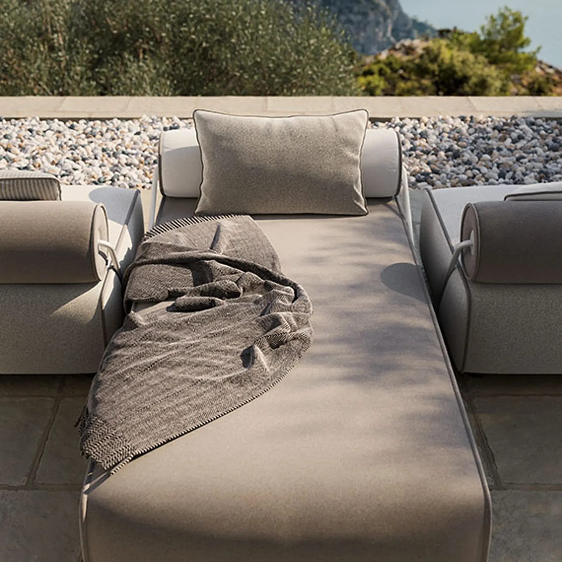 A modern outdoor chaise lounge with a grey pillow and throw blanket on a stone patio overlooking the sea and green hills.