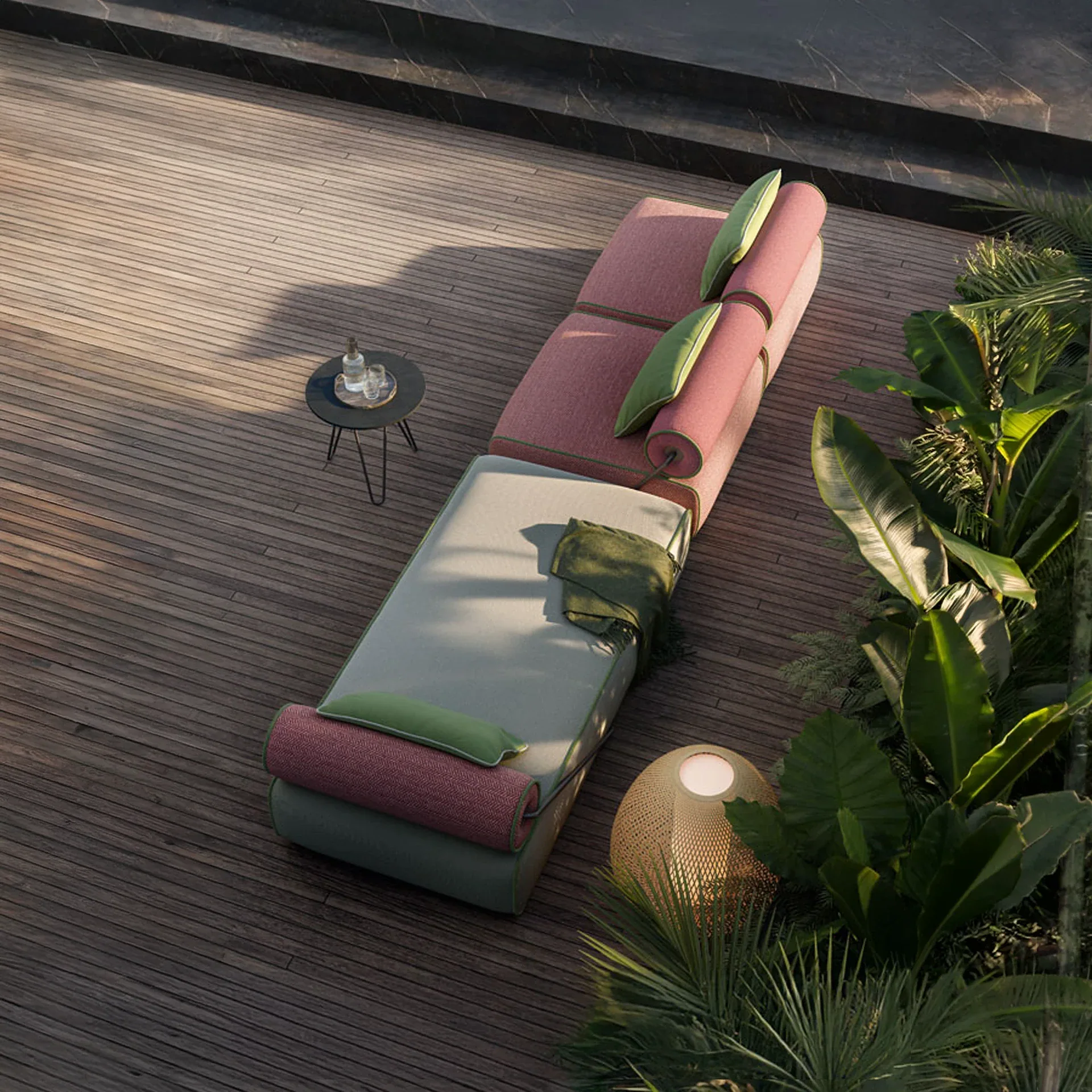 Overhead view of a modern outdoor lounge setup on a wooden deck, featuring a long, modular daybed with pink and grey cushions, green pillows, a side table with drinks, and a woven lamp amidst tropical plants.