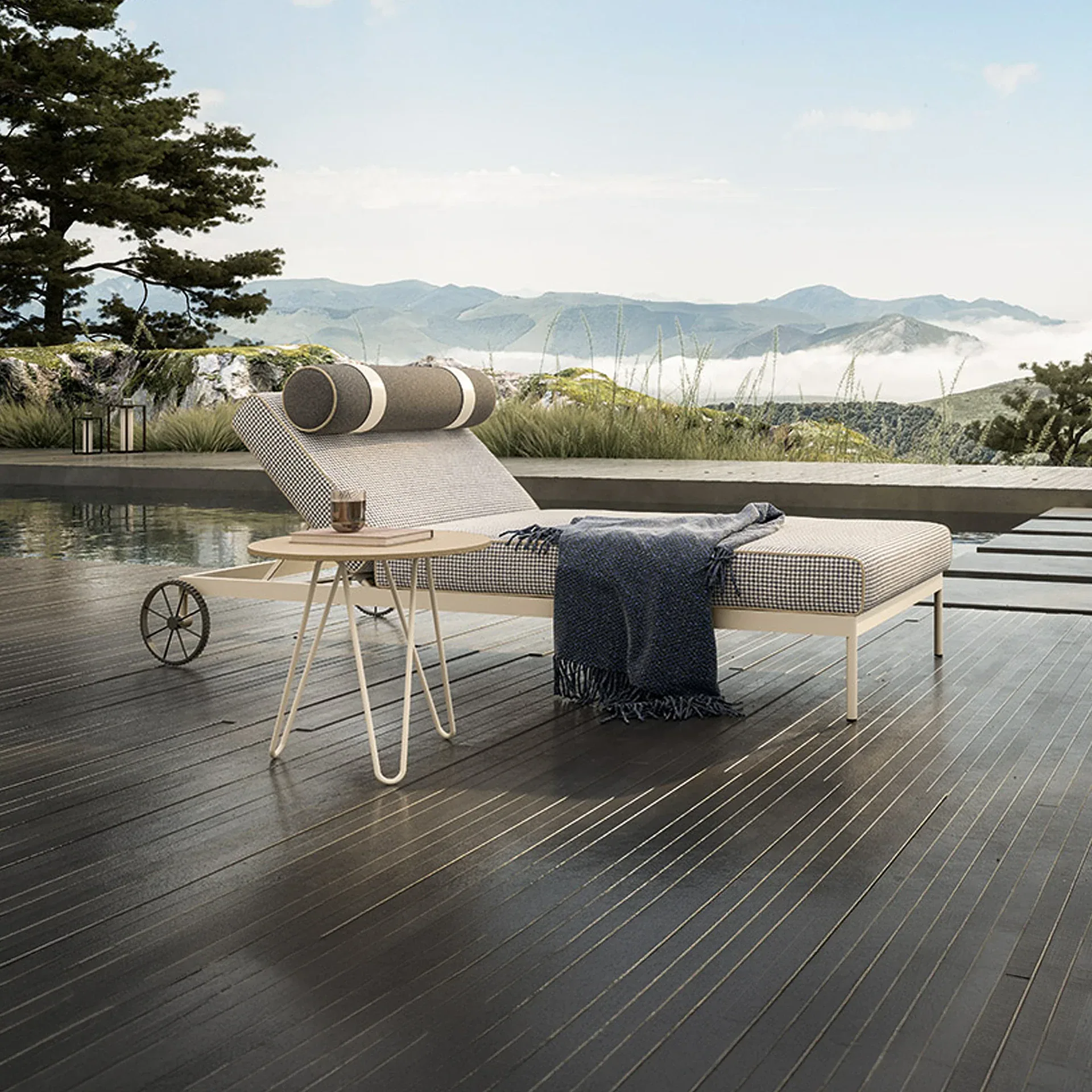 An outdoor lounge chair with a houndstooth cushion and blue blanket on a dark wooden deck next to a pool, overlooking misty mountains.