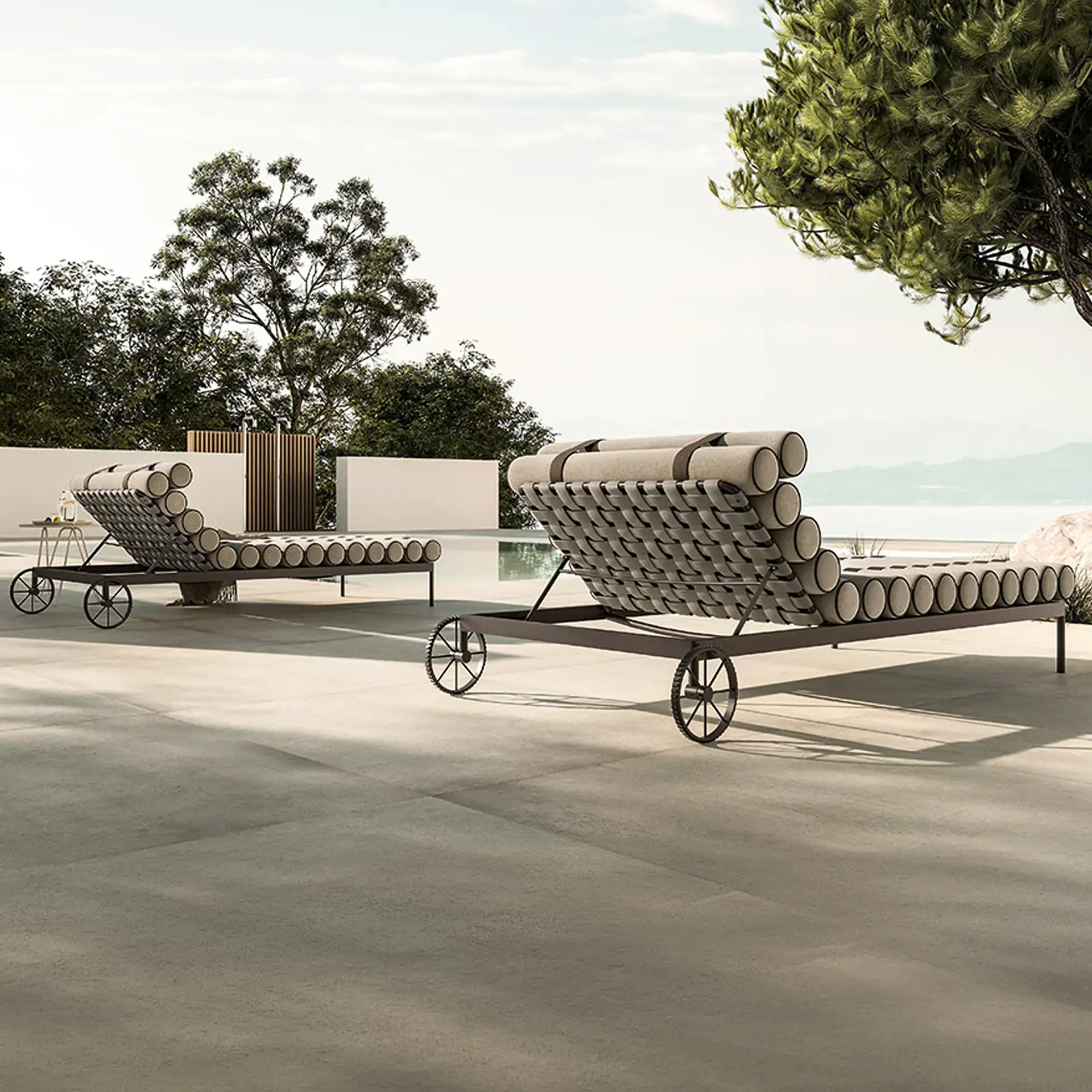 Two modern wheeled lounge chairs with cylindrical cushions on a patio beside an infinity pool, overlooking a sea and mountains under a bright sky.