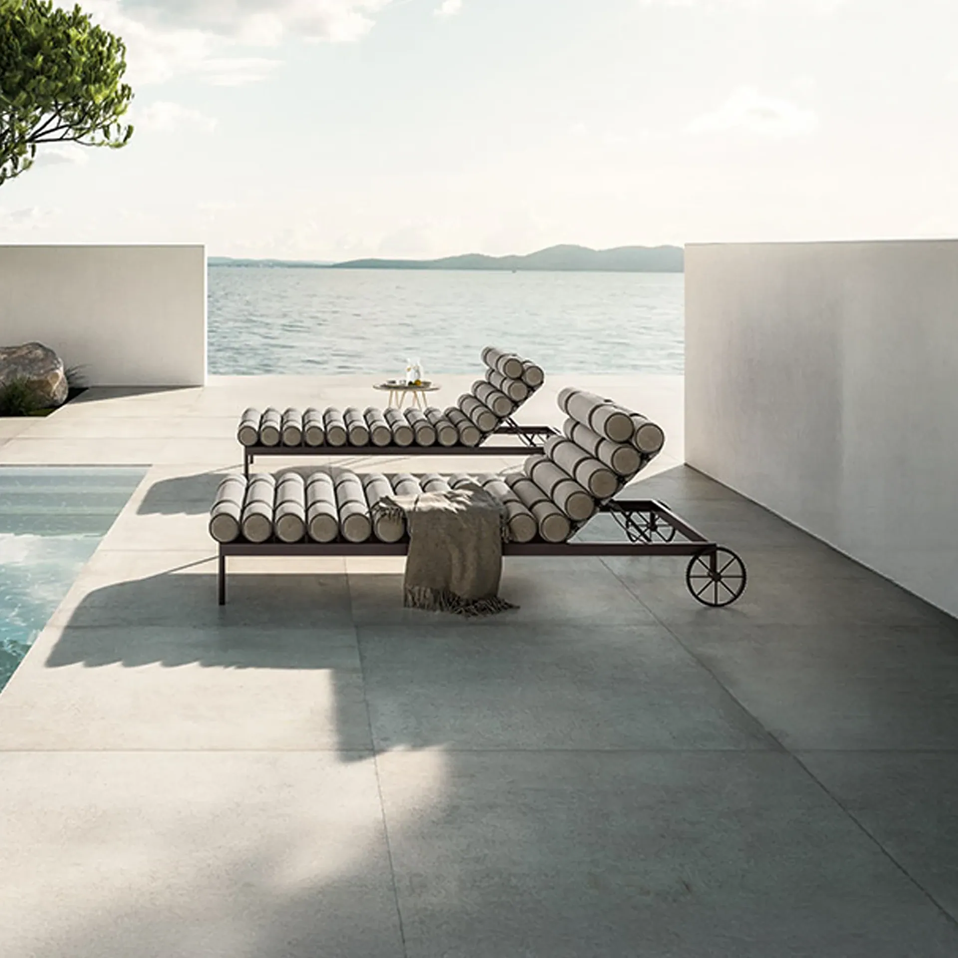 Two modern lounge chairs with cylindrical cushions on a tiled patio overlooking a calm sea and distant hills, with a swimming pool to the side.