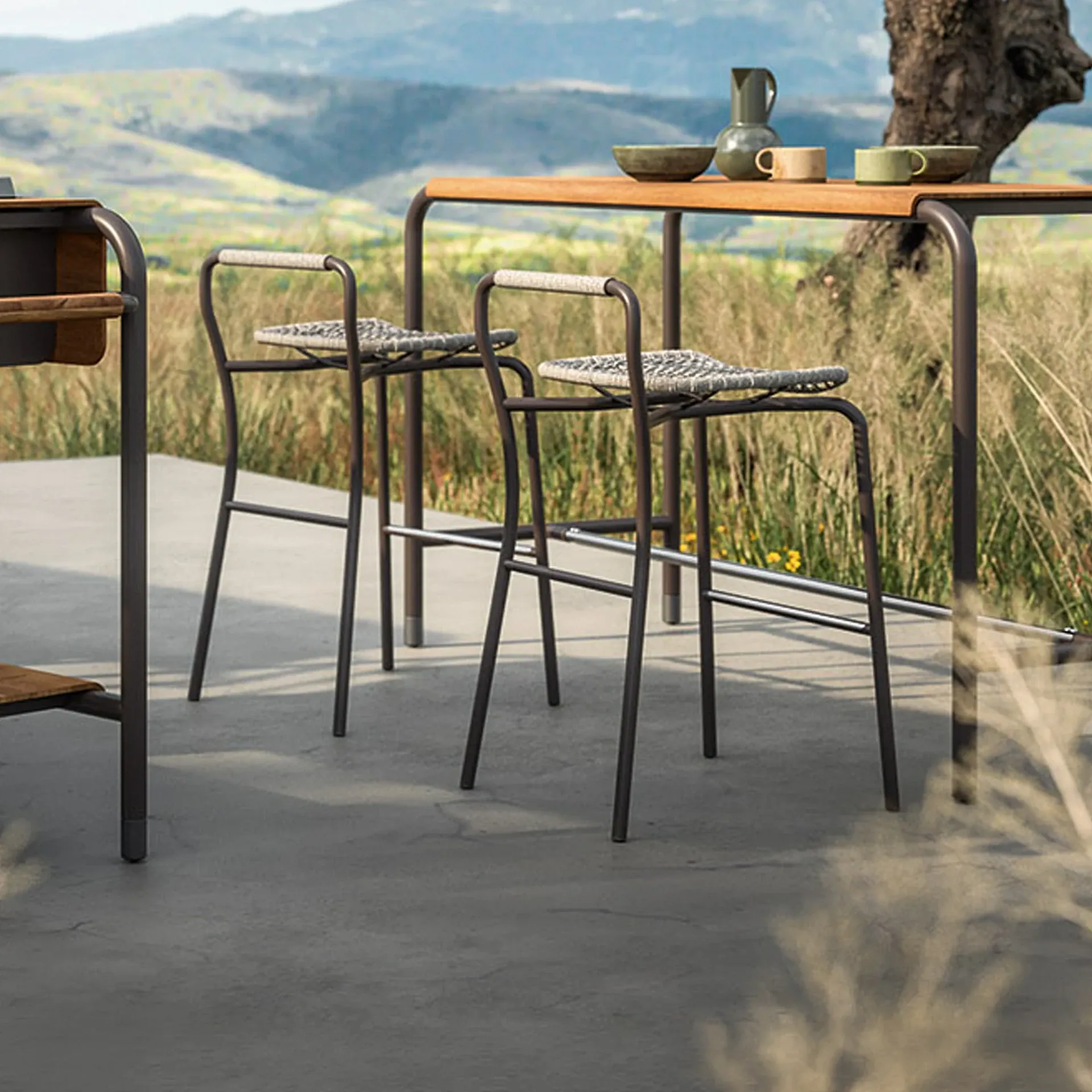 Two modern bar stools and a wooden-top bar table on a concrete patio, overlooking grassy hills and a tree.