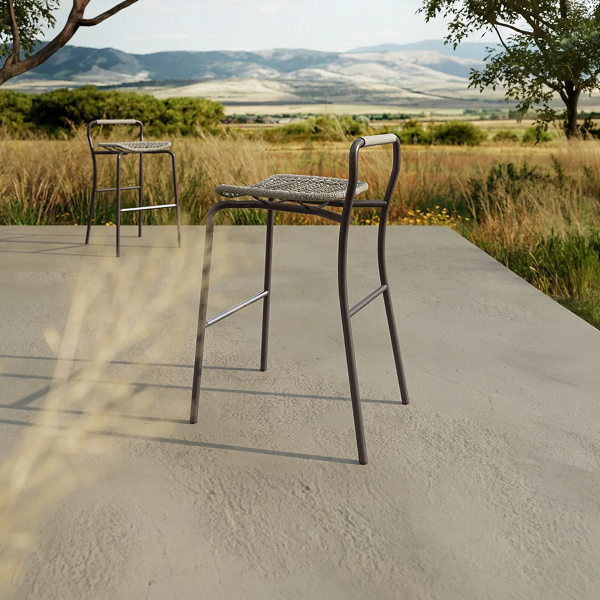 Two modern bar stools with metal frames and woven seats on a concrete patio overlooking a vast landscape of dry grasses and distant mountains.