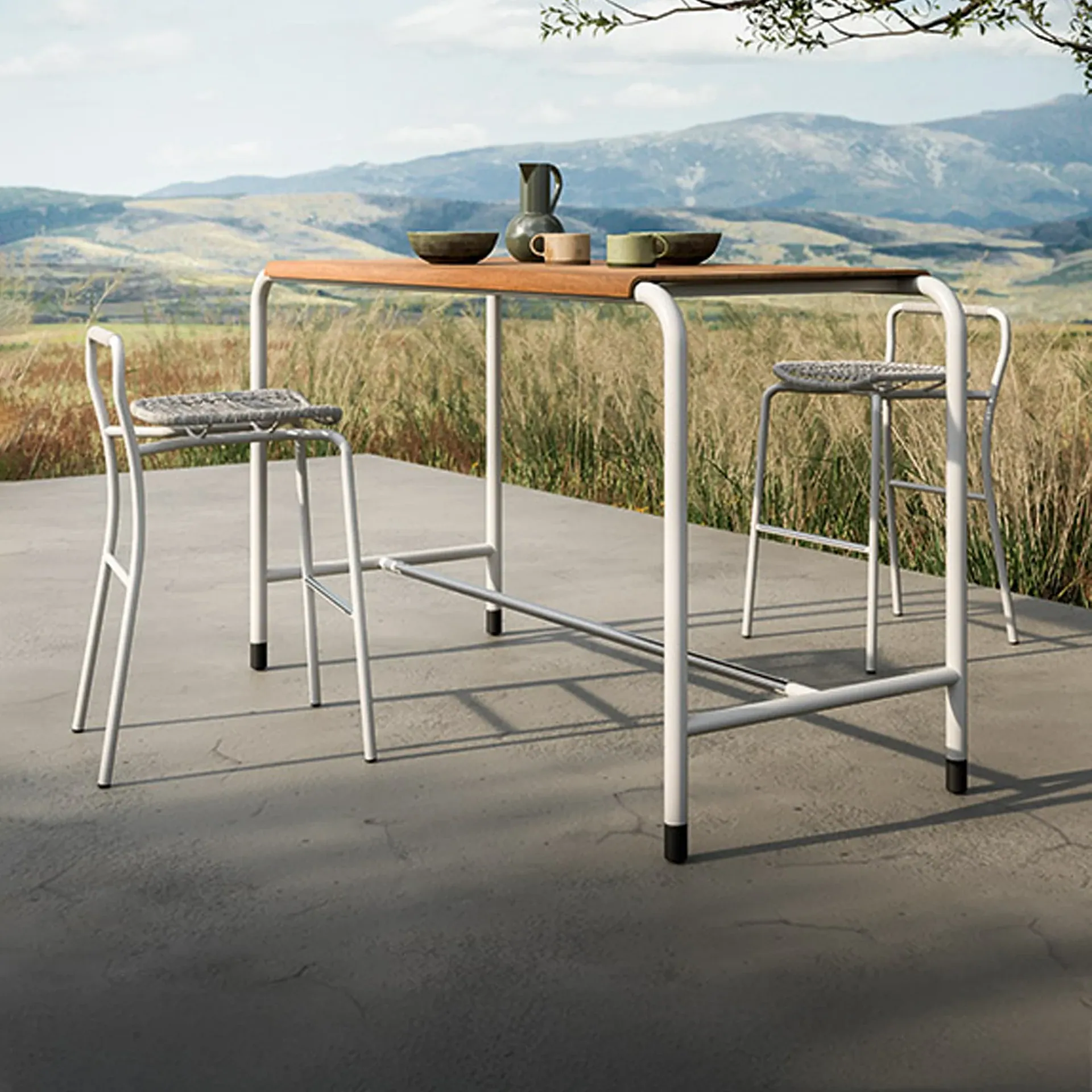 An outdoor bar table with a wooden top and white metal frame, accompanied by two white bar stools with woven seats, set on a concrete patio overlooking a scenic mountain landscape with dry grasses.