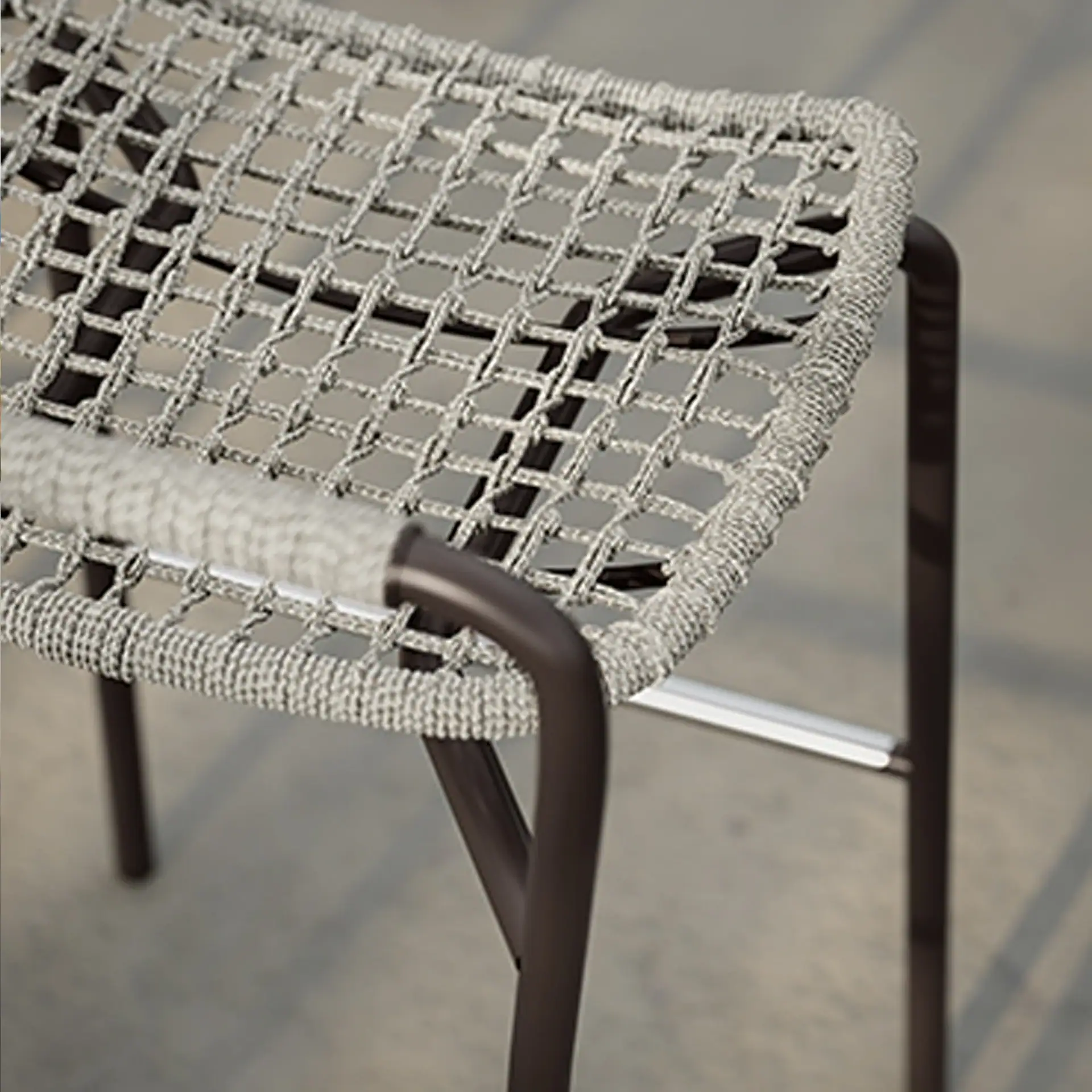 Close-up of a stool with a light grey woven rope seat and dark brown metal legs, on a blurred concrete surface.