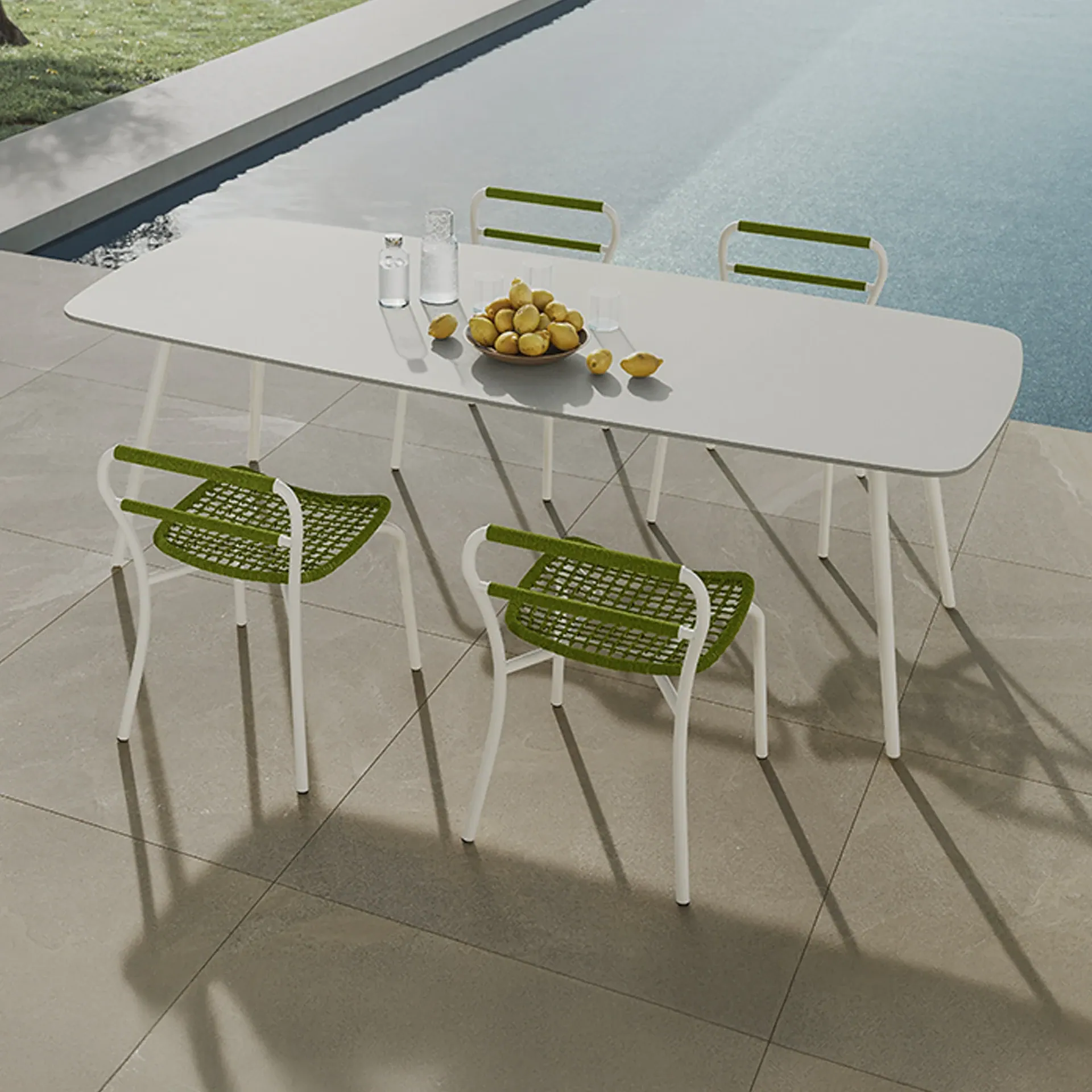 An outdoor dining table with a bowl of lemons and drinks, surrounded by four modern white and green chairs, set on a tiled patio next to a swimming pool.