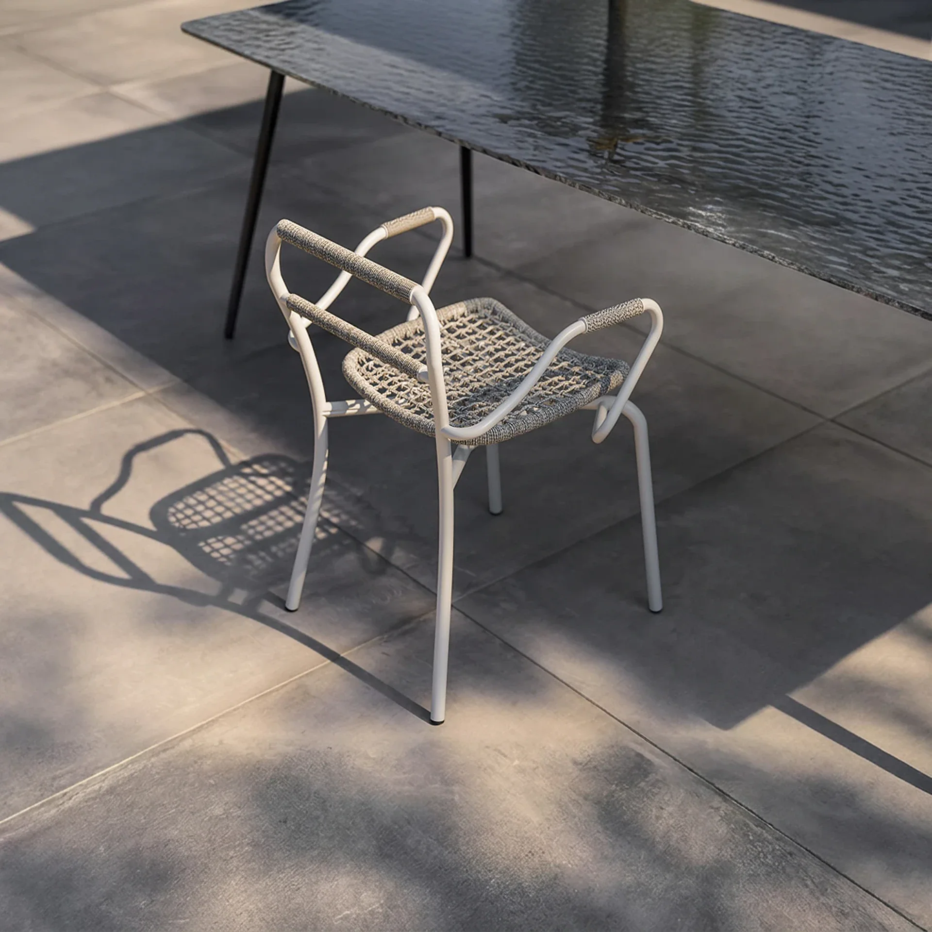 A modern white metal chair with a woven grey seat on a grey tiled patio, next to a dark textured table, with strong shadows cast by sunlight.