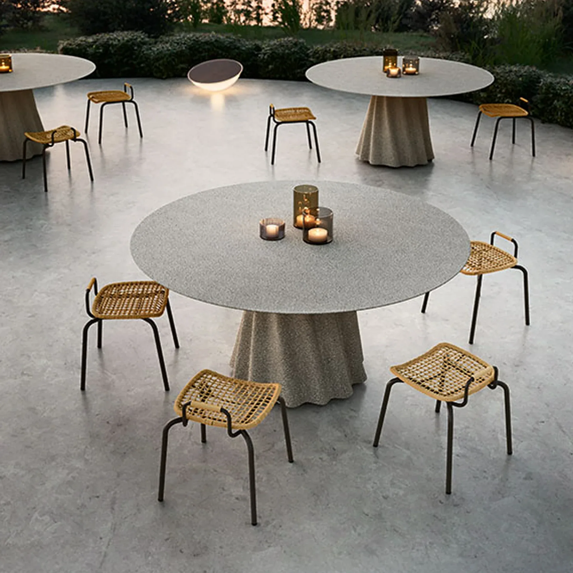Outdoor patio with three round tables, wicker stools, and lit candles, surrounded by greenery and a crescent-shaped light.