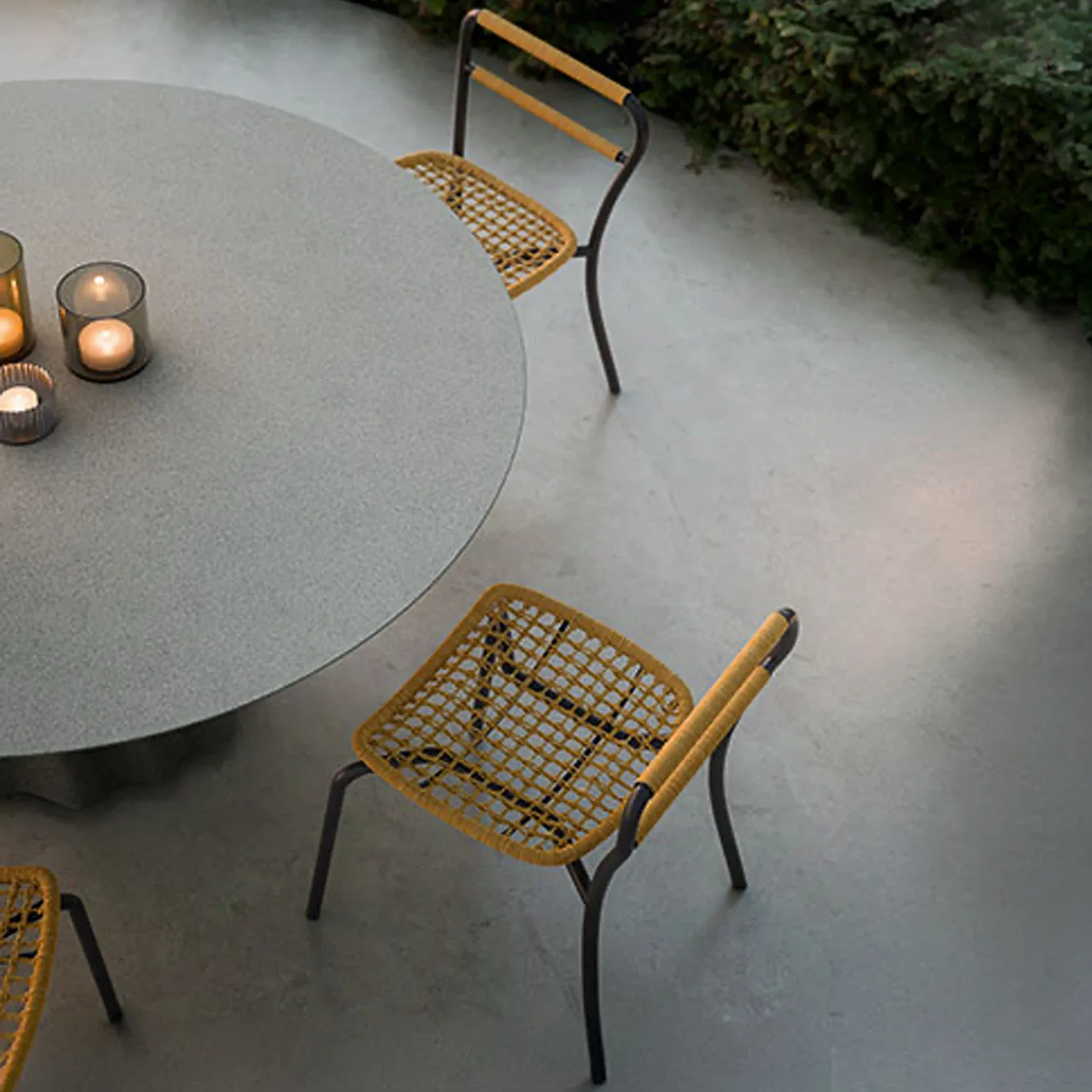 Overhead view of a round grey outdoor table with three yellow-orange woven chairs and lit candles on a concrete patio next to green foliage.