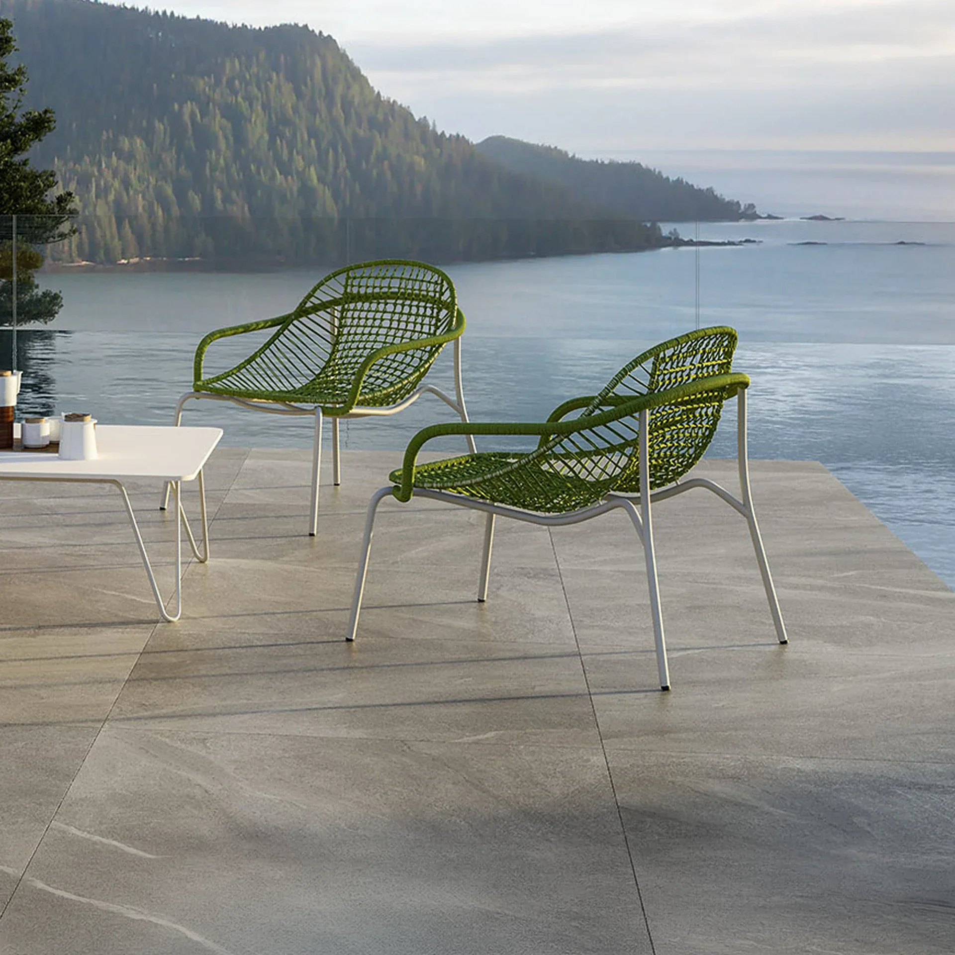 Two modern green outdoor chairs and a white coffee table on a patio overlooking a vast ocean and distant forested mountains.