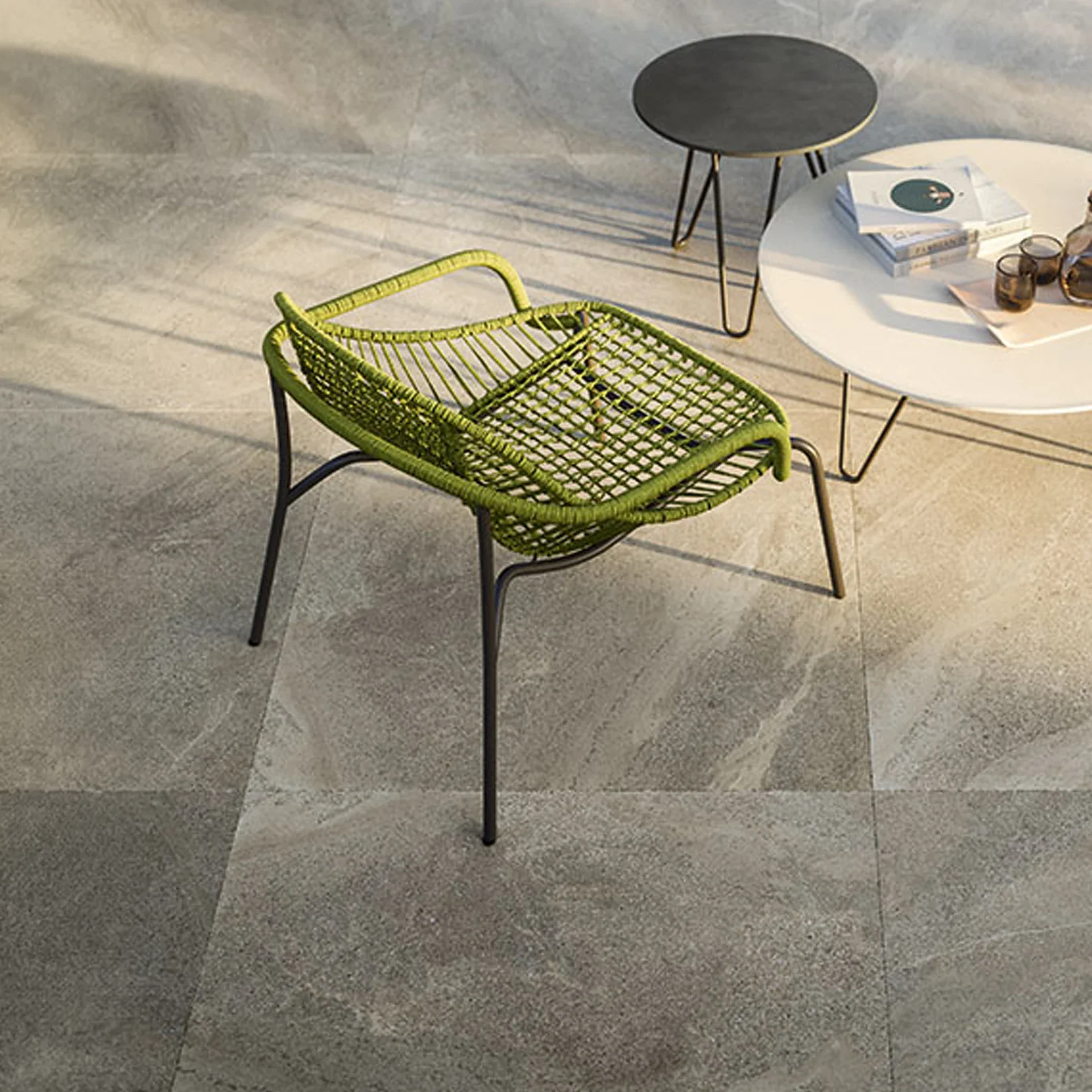 An outdoor patio featuring a lime green woven chair, a white round table with books and drinks, and a smaller dark grey round table, all on grey tiled flooring.