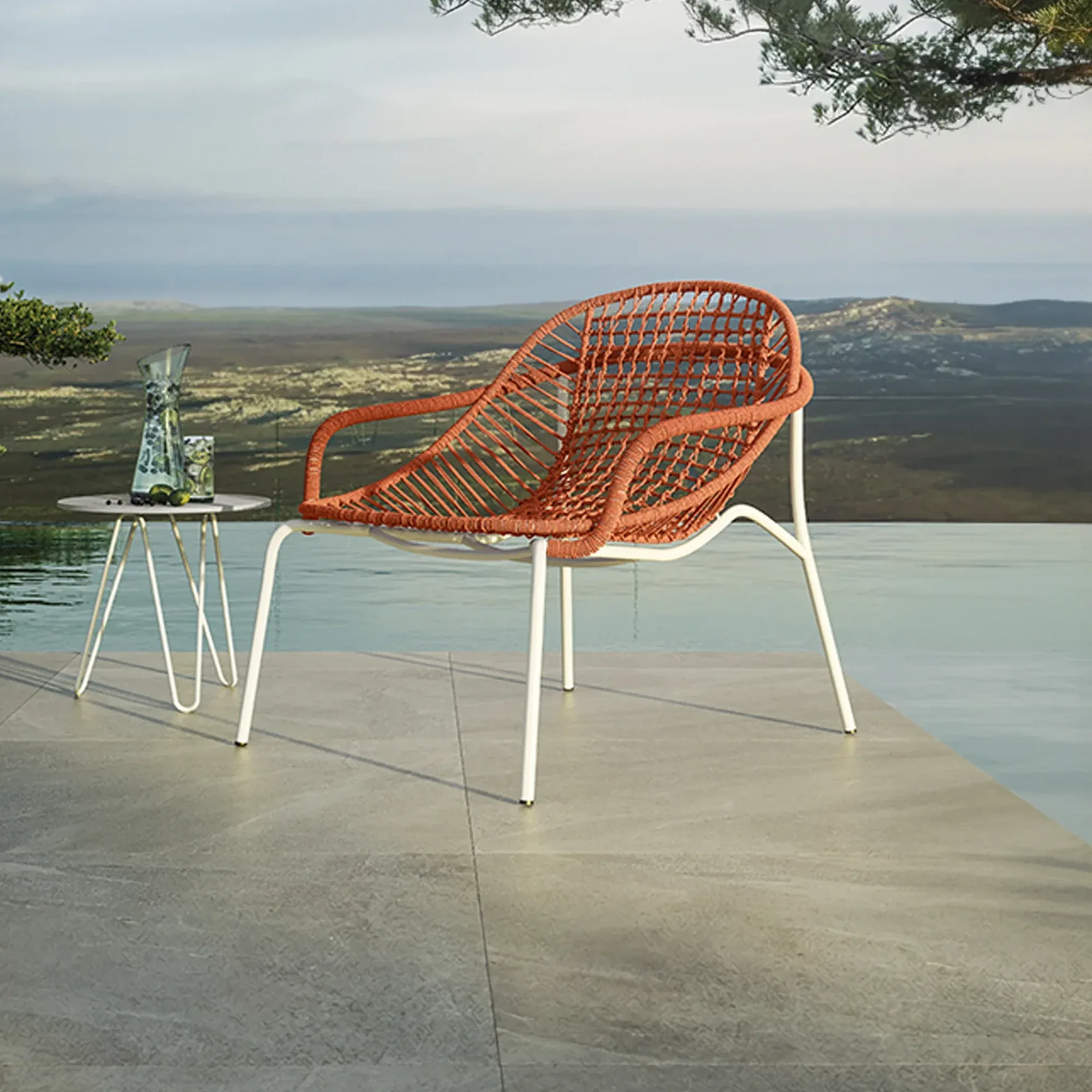 A modern orange woven outdoor chair and a small side table with a pitcher and glass on a grey tiled patio next to an infinity pool, overlooking a distant landscape under a cloudy sky.