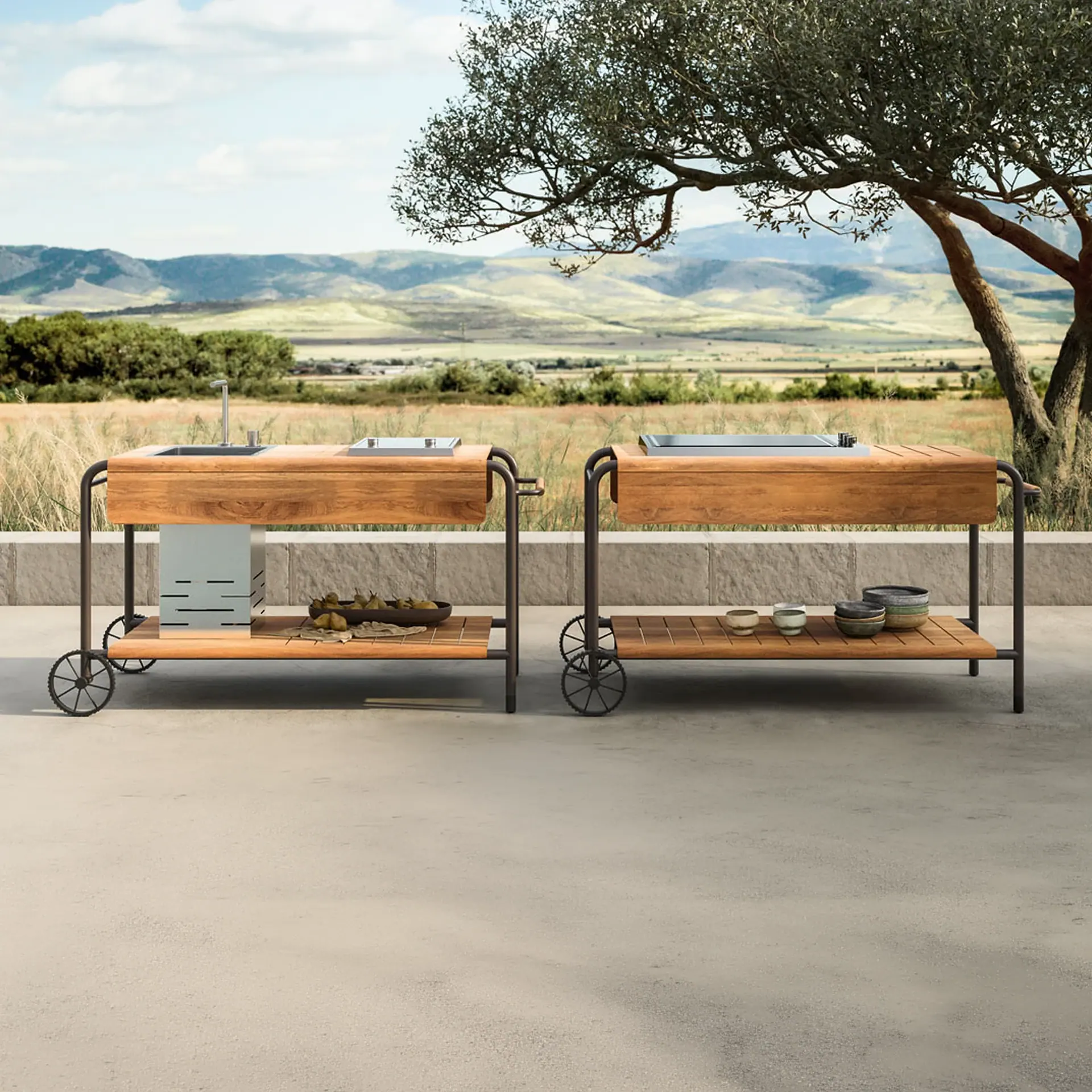 Two modern wooden outdoor kitchen carts with metal frames and wheels on a patio, set against a backdrop of rolling hills and a large tree.