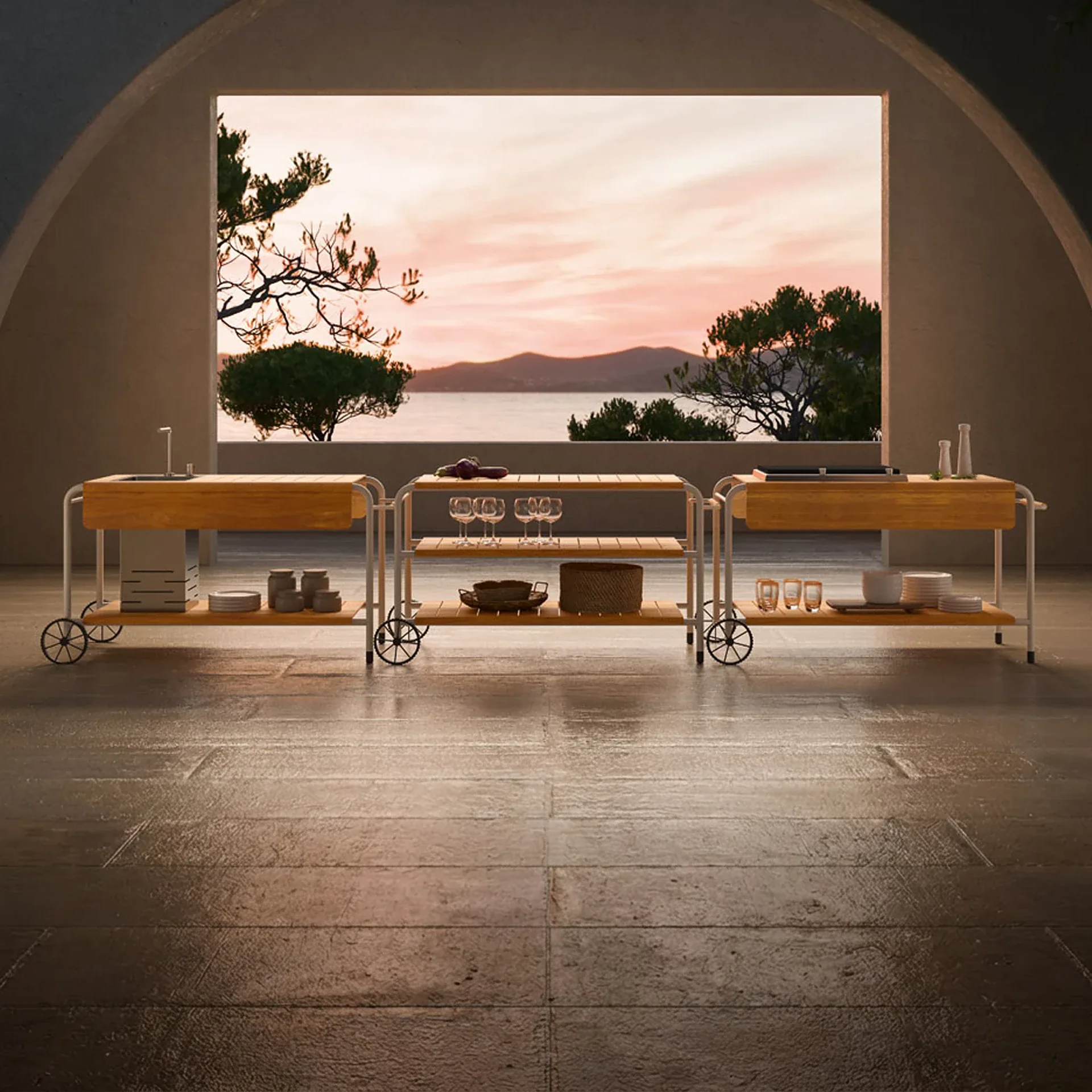 Three mobile kitchen carts with wooden tops and white frames arranged on a dark tiled patio, overlooking a sunset view of the sea, mountains, and trees through a large rectangular opening.