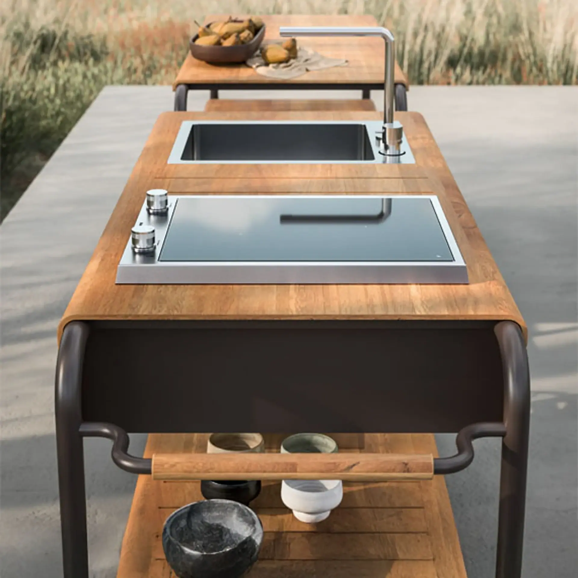 A modern outdoor kitchen unit with a wooden countertop, stainless steel sink, sleek faucet, and black glass cooktop. A lower wooden shelf holds decorative bowls. The unit is set outdoors with blurred greenery in the background.