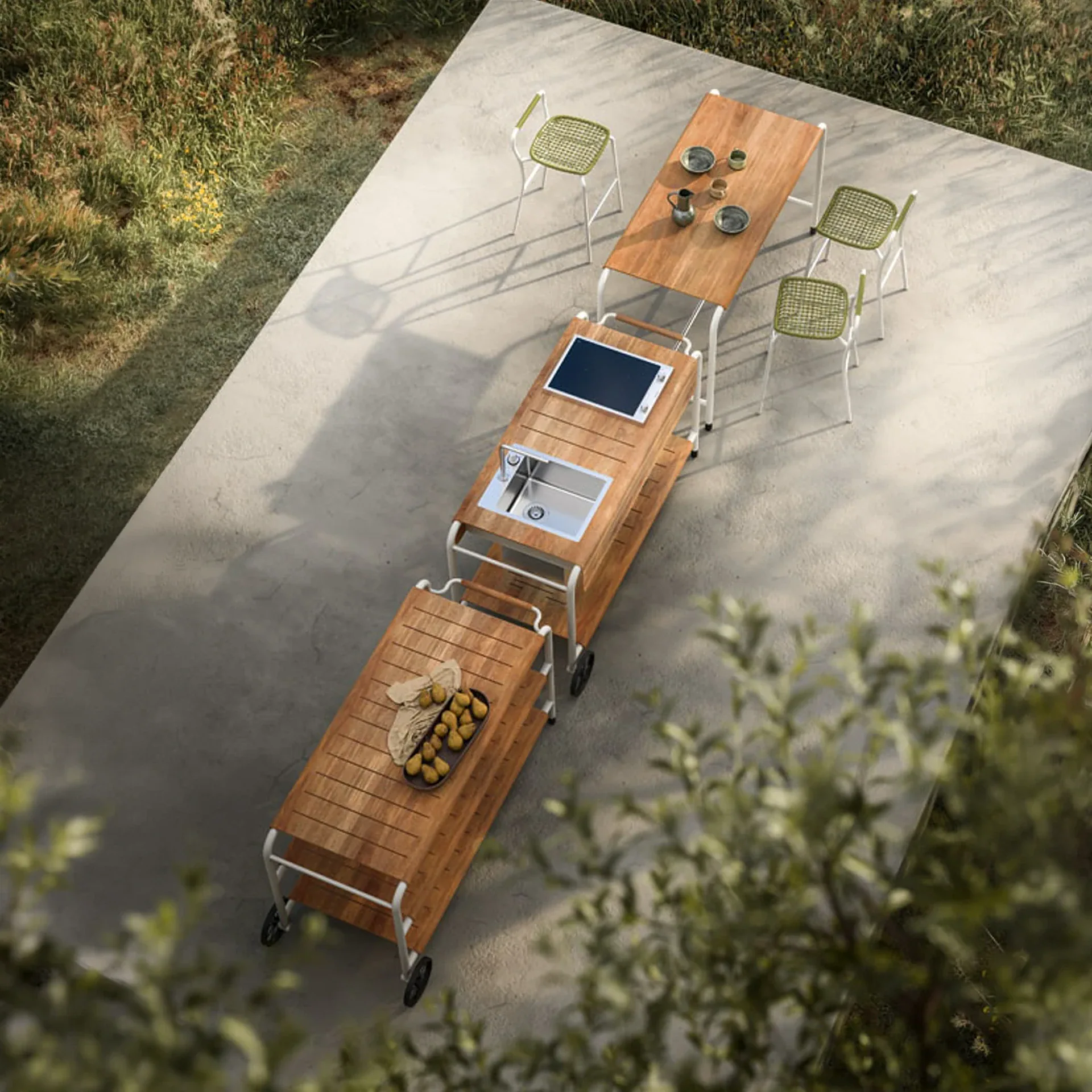 An overhead view of a modular outdoor kitchen on a concrete patio, featuring a dining table with chairs, a sink unit, and a prep cart with pears, surrounded by green foliage.
