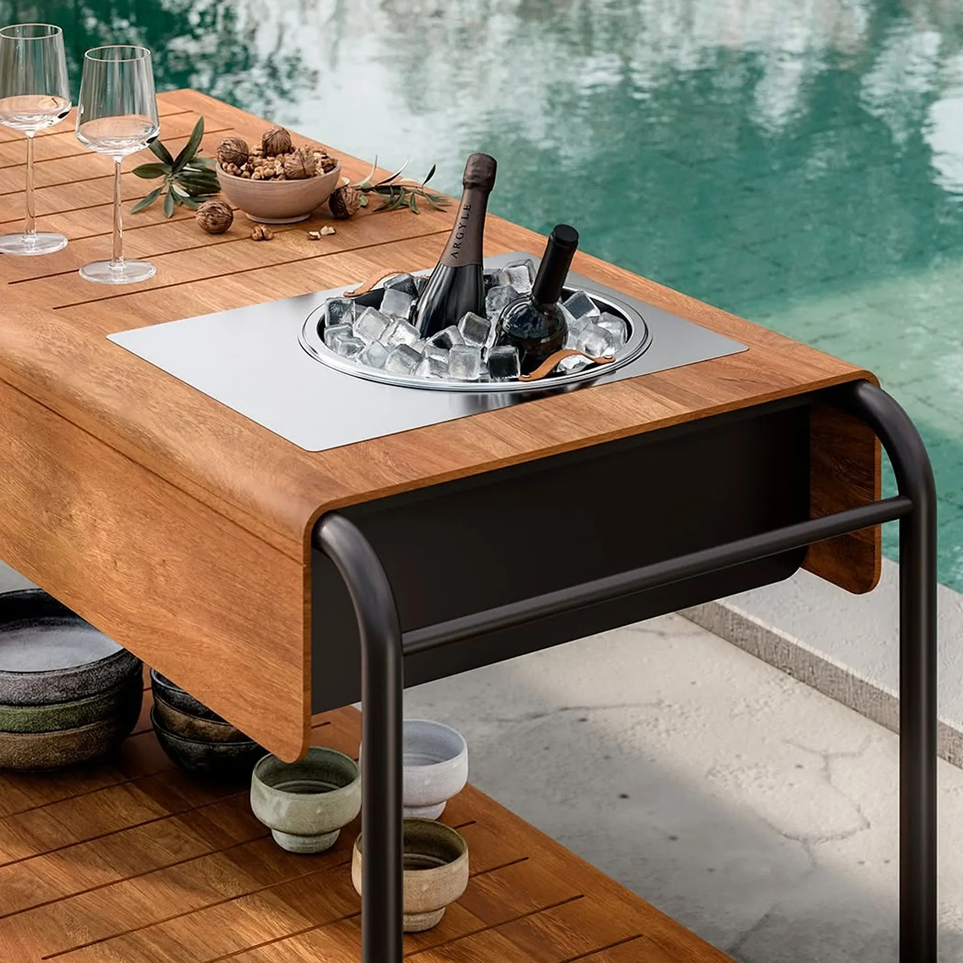 A wooden outdoor serving cart with an integrated stainless steel ice bucket holding two bottles, two wine glasses, and a bowl of walnuts, positioned next to a swimming pool.