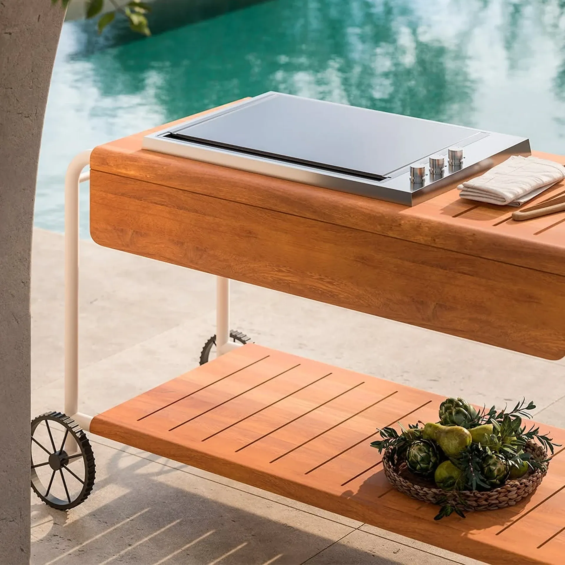 A modern outdoor grill cart with a stainless steel griddle, wooden shelves, and a basket of produce, set beside a swimming pool.