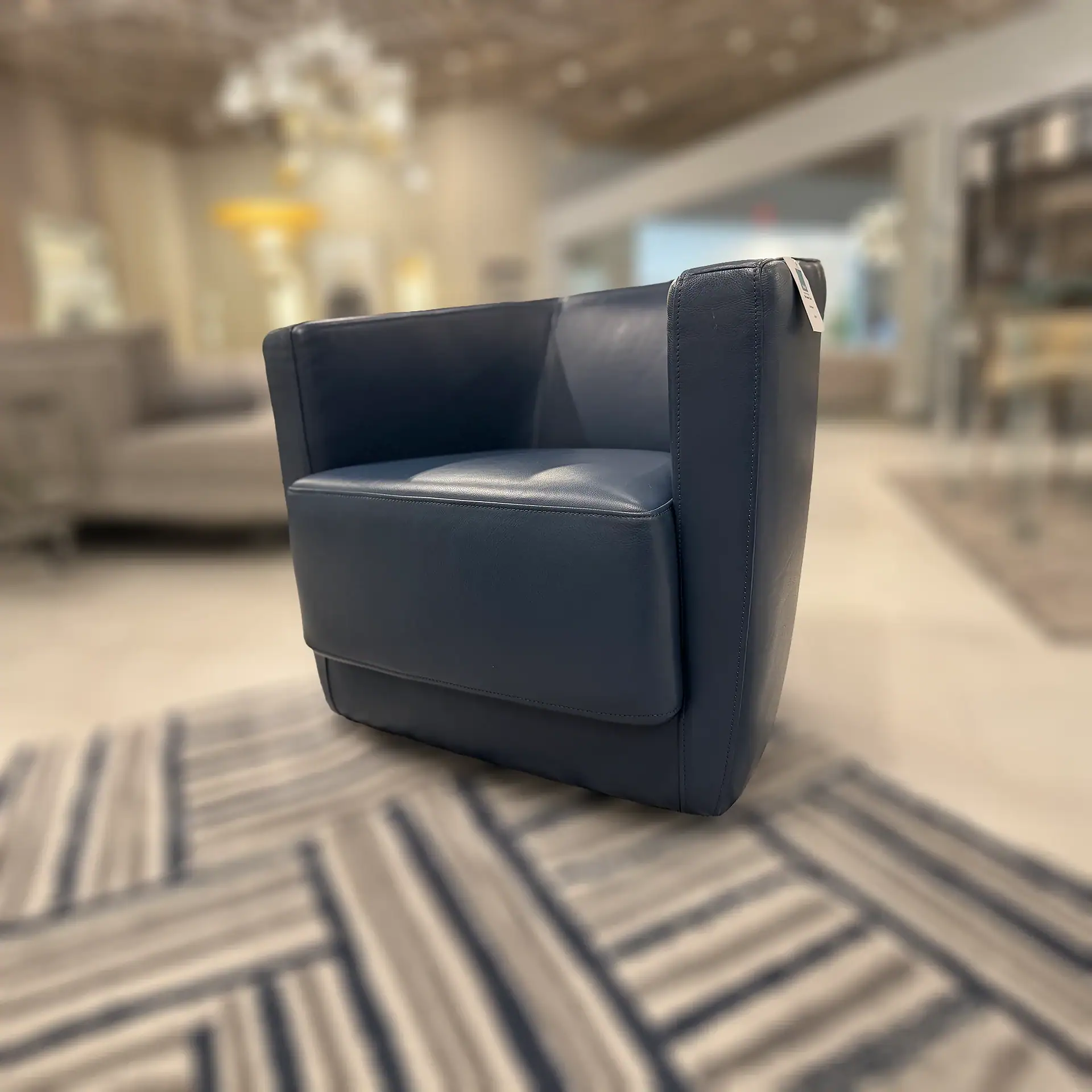 A modern navy blue leather armchair with a boxy design, sitting on a geometric patterned rug in a blurred showroom background.