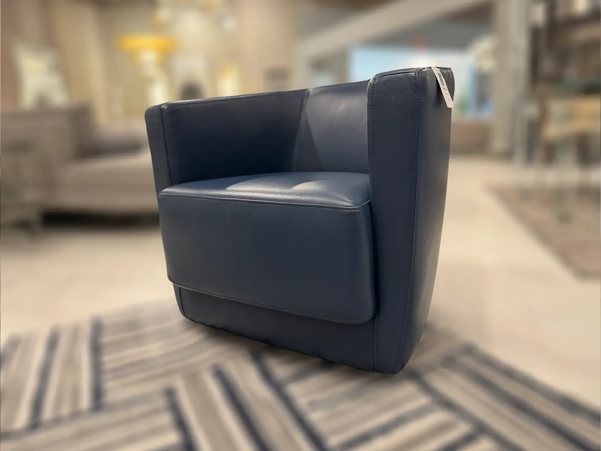 A dark blue modern armchair with visible stitching sits on a striped rug in a blurred showroom setting.