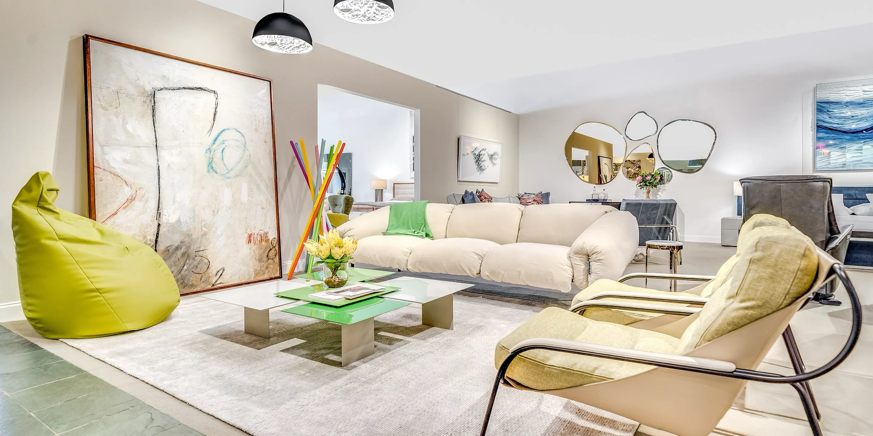 showroom-vignette-apr-24-living-room A modern living room with a cream sofa, green bean bag, abstract art, and unique mirrors, featuring a colorful and contemporary design.