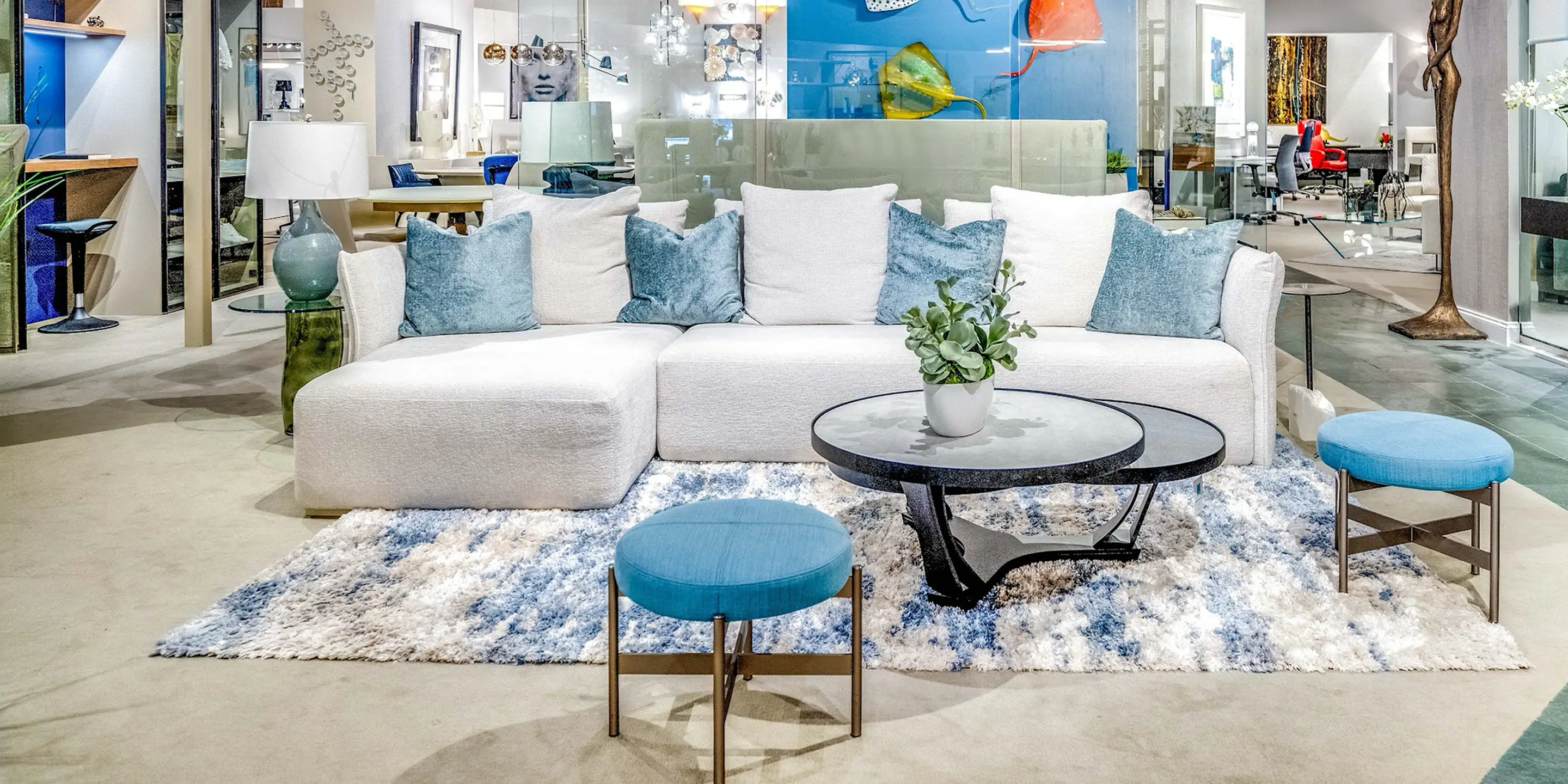 showroom-vignette-aug-23-living-room-2 A modern white sectional sofa with blue throw pillows, a glass coffee table with a plant, and two blue ottomans on an abstract blue and white rug in a showroom.