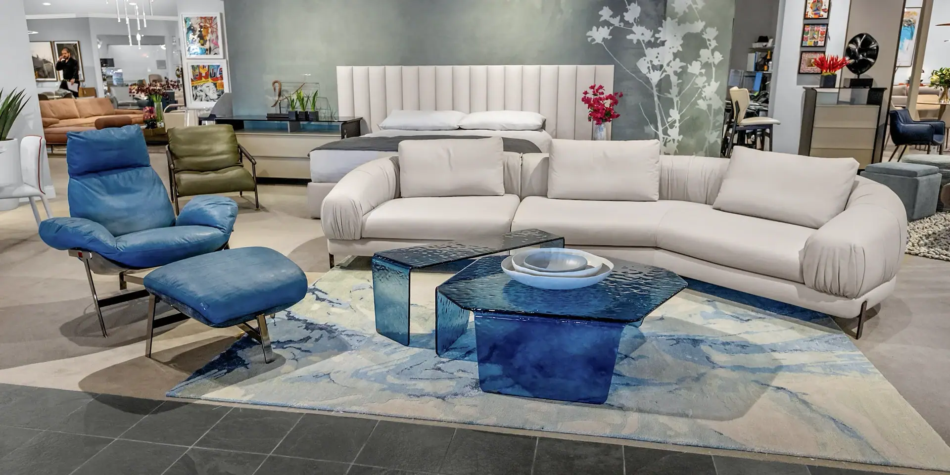 A modern living room display with a cream sectional sofa, blue glass coffee tables, a blue leather armchair and ottoman, and a bed with a cream headboard in the background.