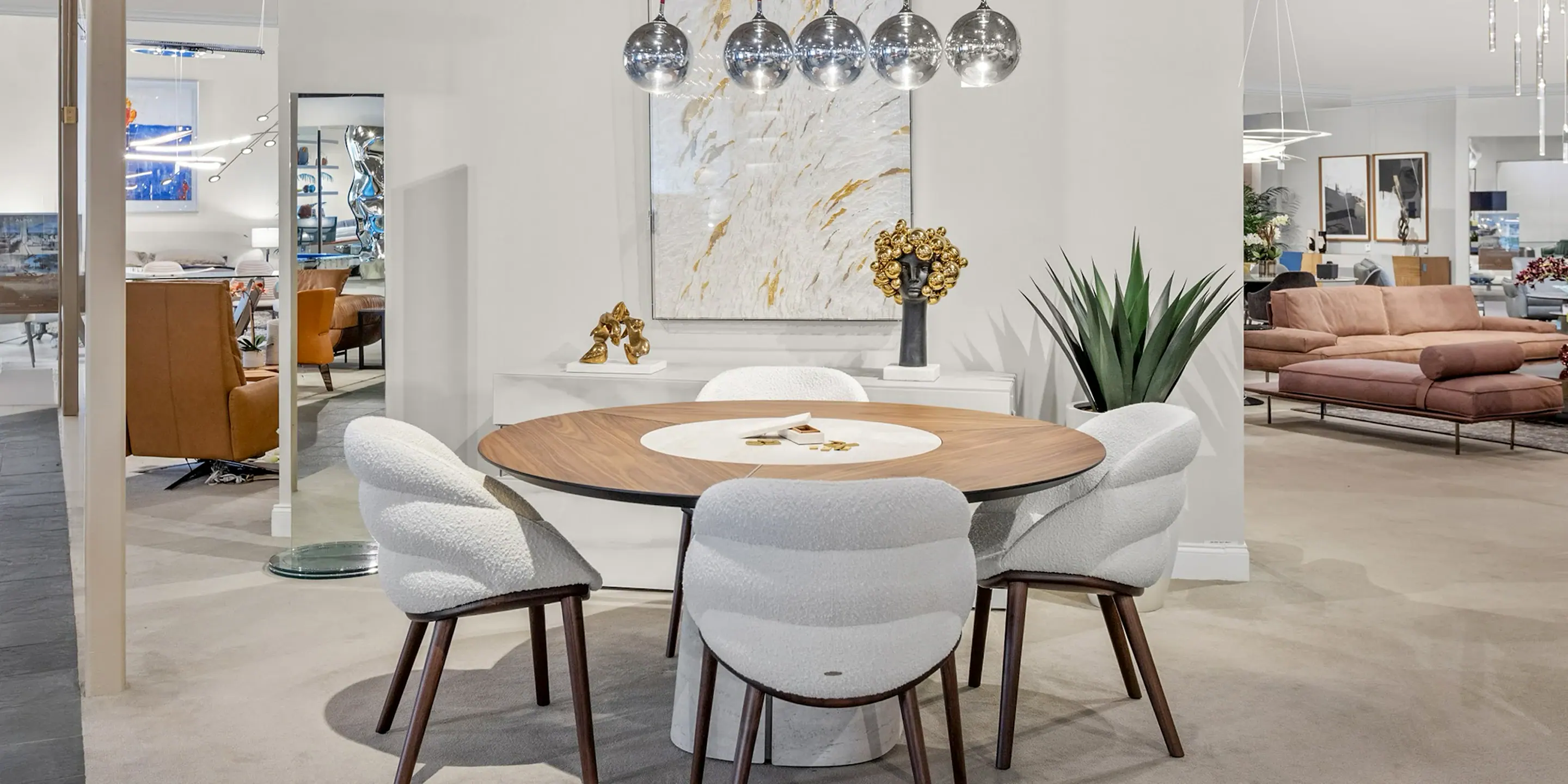 showroom-vignette-feb-25-dining-room-2 A round wooden dining table with a light center insert, surrounded by four white textured chairs with wooden legs, under spherical pendant lights. An abstract gold and white painting hangs on the wall behind, flanked by modern sculptures and a tall plant. Other furniture displays are visible in the background.