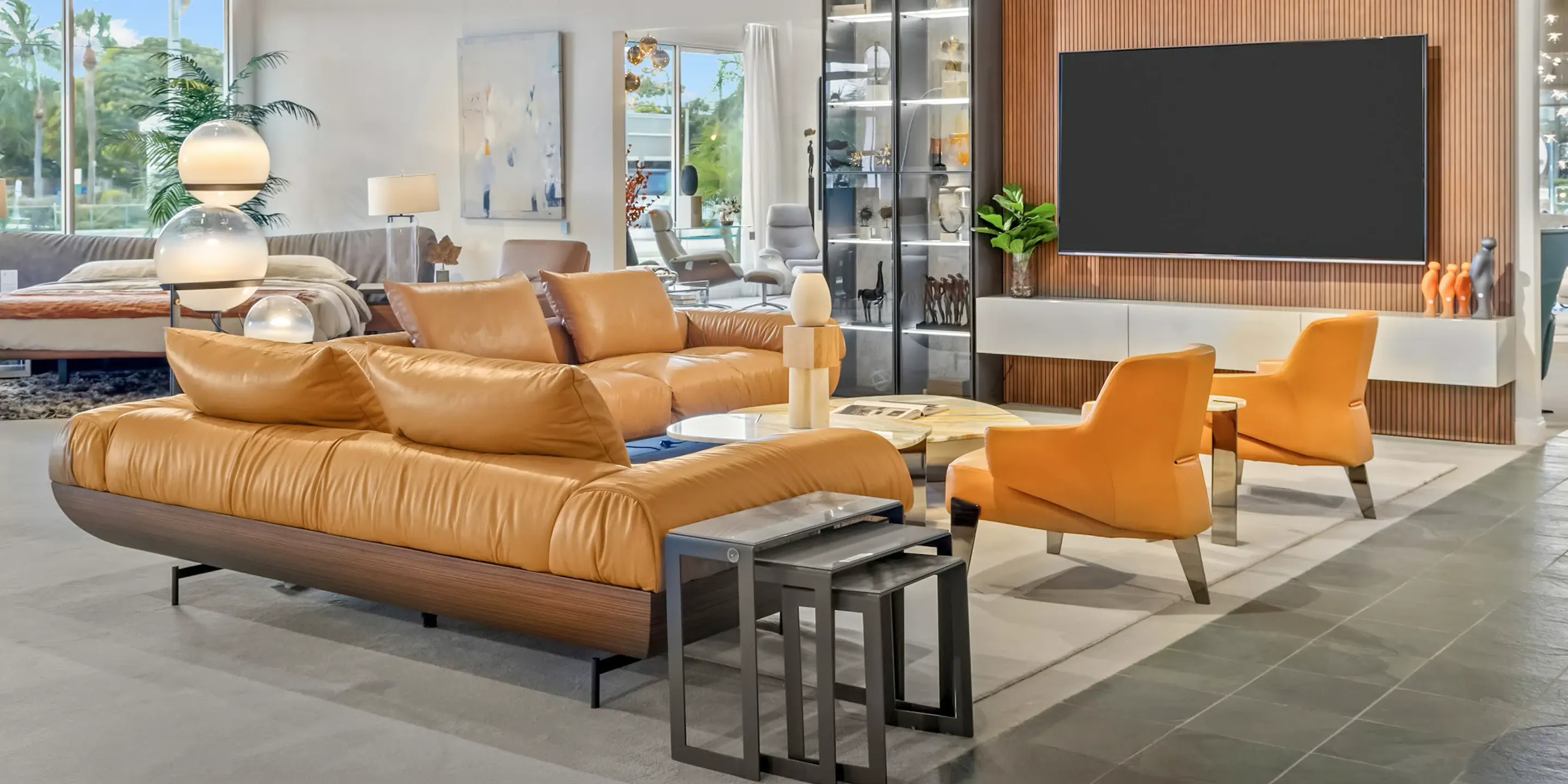 showroom-vignette-oct-24-living-room A modern living room with a tan leather L-shaped sofa, two orange armchairs, marble coffee tables, and a large TV mounted on a wooden slatted wall.